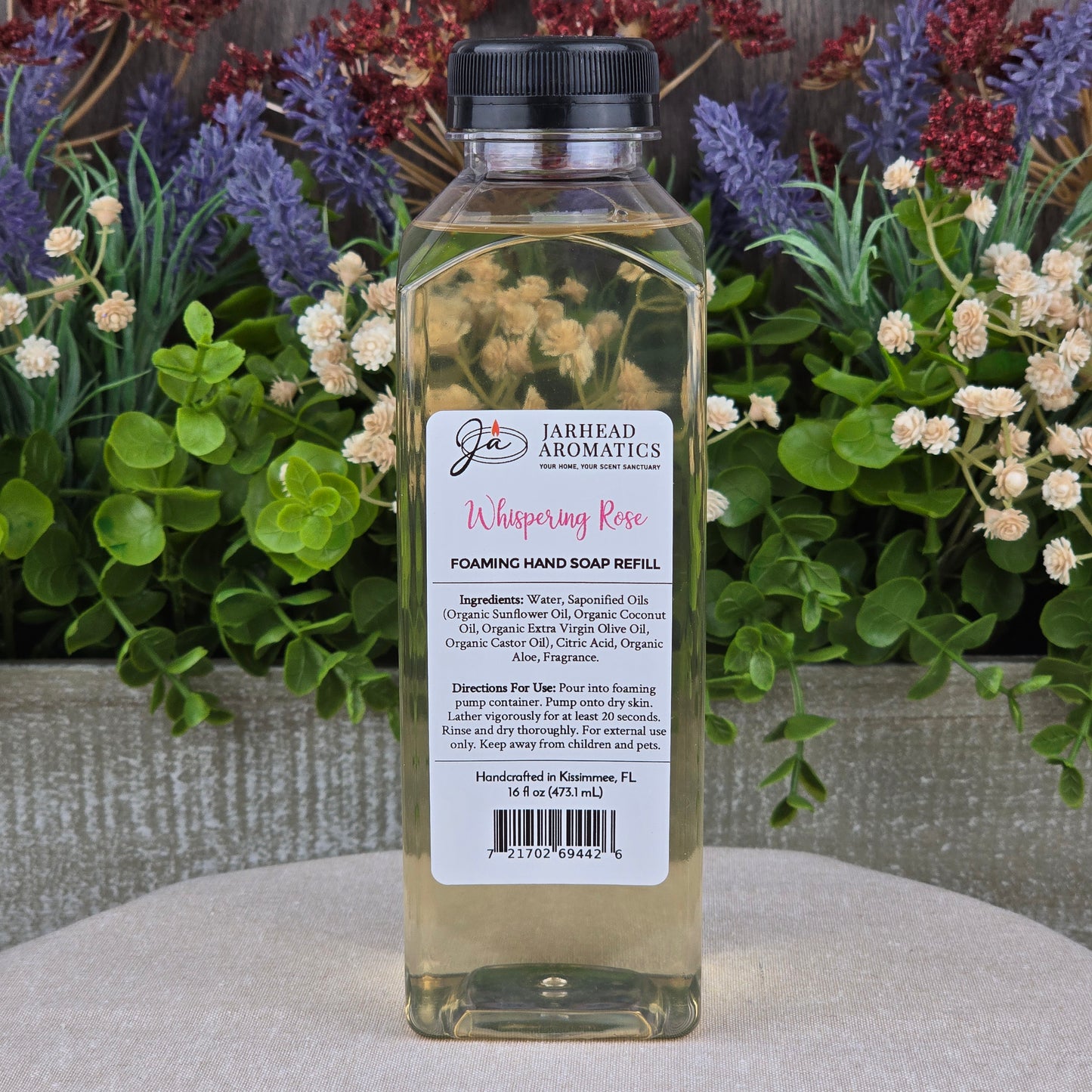 A clear bottle of Jarhead Aromatics Whispering Rose Foaming Hand Soap rests on a gray surface. Its label lists ingredients and directions, with green plants and colorful flowers in the background for a touch of botanical luxury.