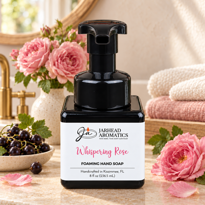 A square bottle of Jarhead Aromatics Whispering Rose Handmade Foaming Hand Soap with a black pump sits on a counter next to a small bowl of black currants and pink roses.