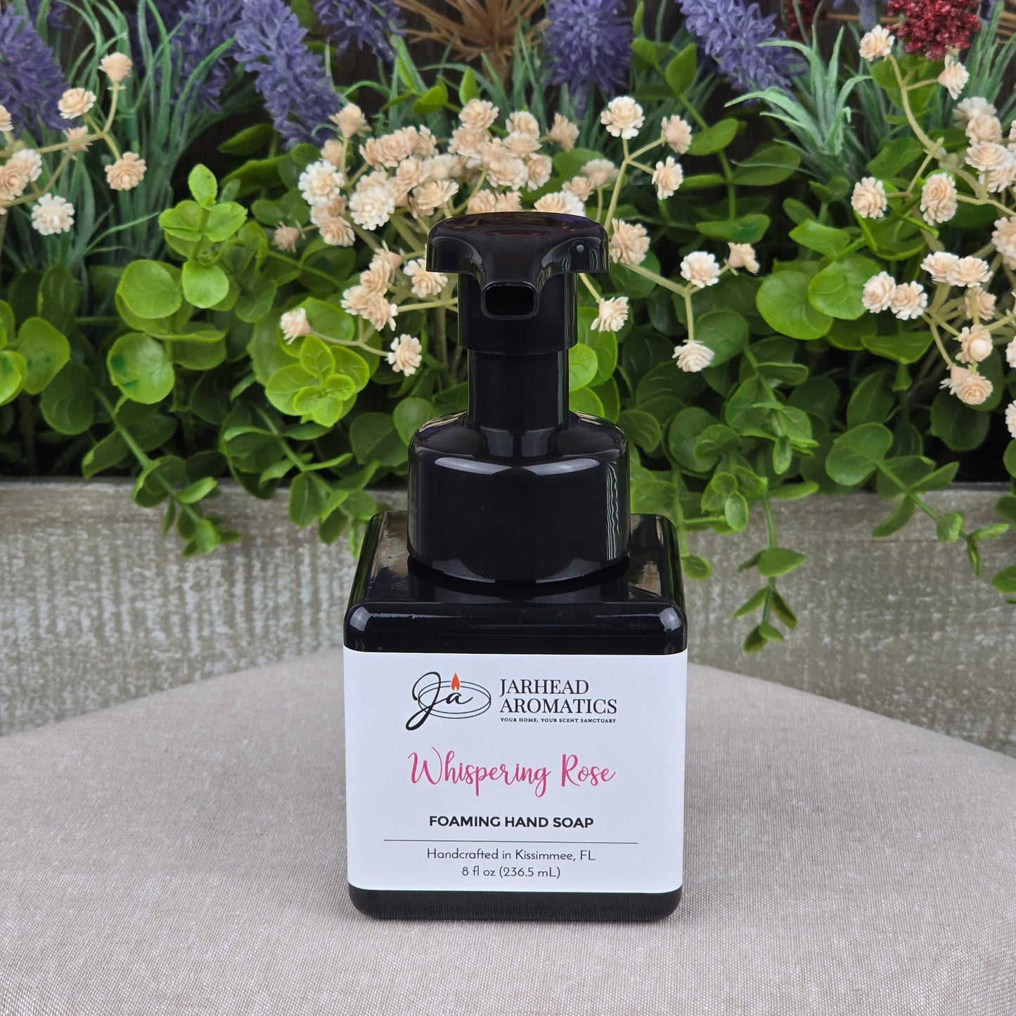 A bottle of Jarhead Aromatics Whispering Rose Foaming Hand Soap rests on a gray surface, surrounded by artificial flowers and green foliage.