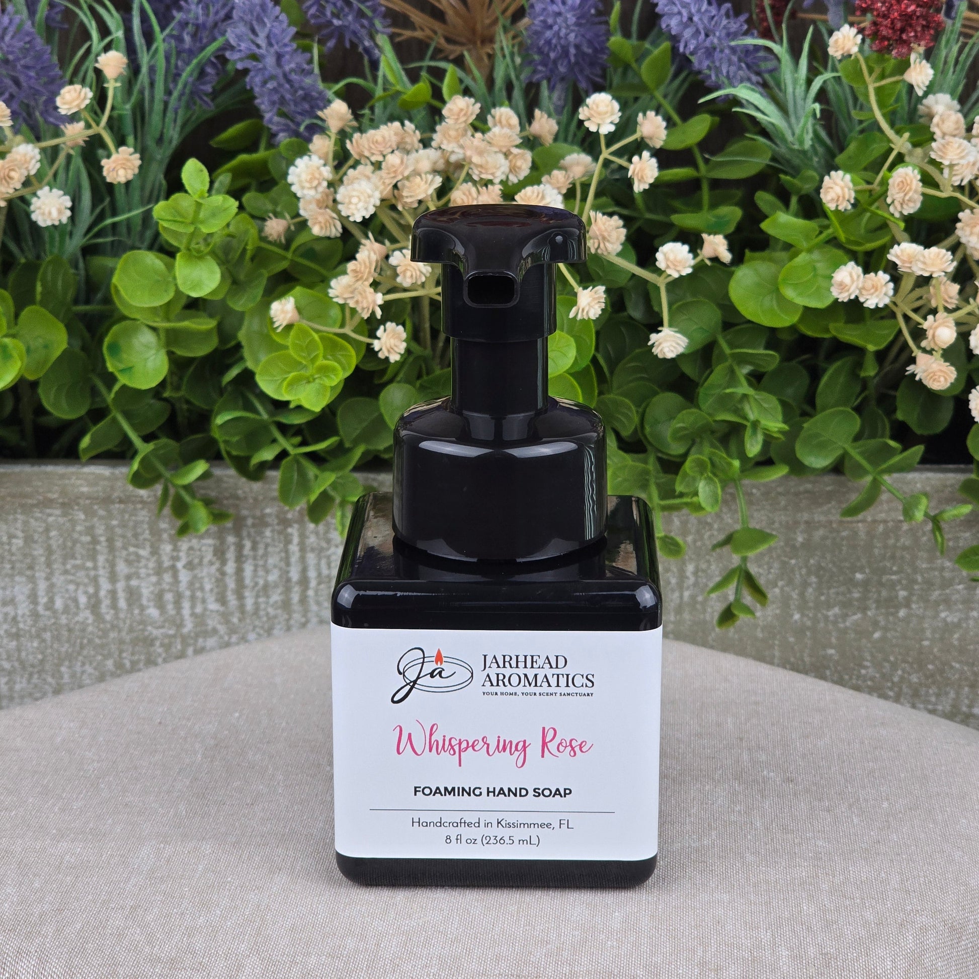 A bottle of Jarhead Aromatics Whispering Rose Foaming Hand Soap rests on a gray surface, surrounded by artificial flowers and green foliage.