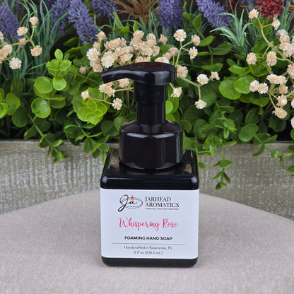 A bottle of Jarhead Aromatics Whispering Rose Foaming Hand Soap rests on a light surface, surrounded by artificial flowers and greenery.
