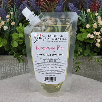 A clear pouch of Jarhead Aromatics Whispering Rose Foaming Hand Soap sits on a gray surface, with artificial green and purple flowers in the background.
