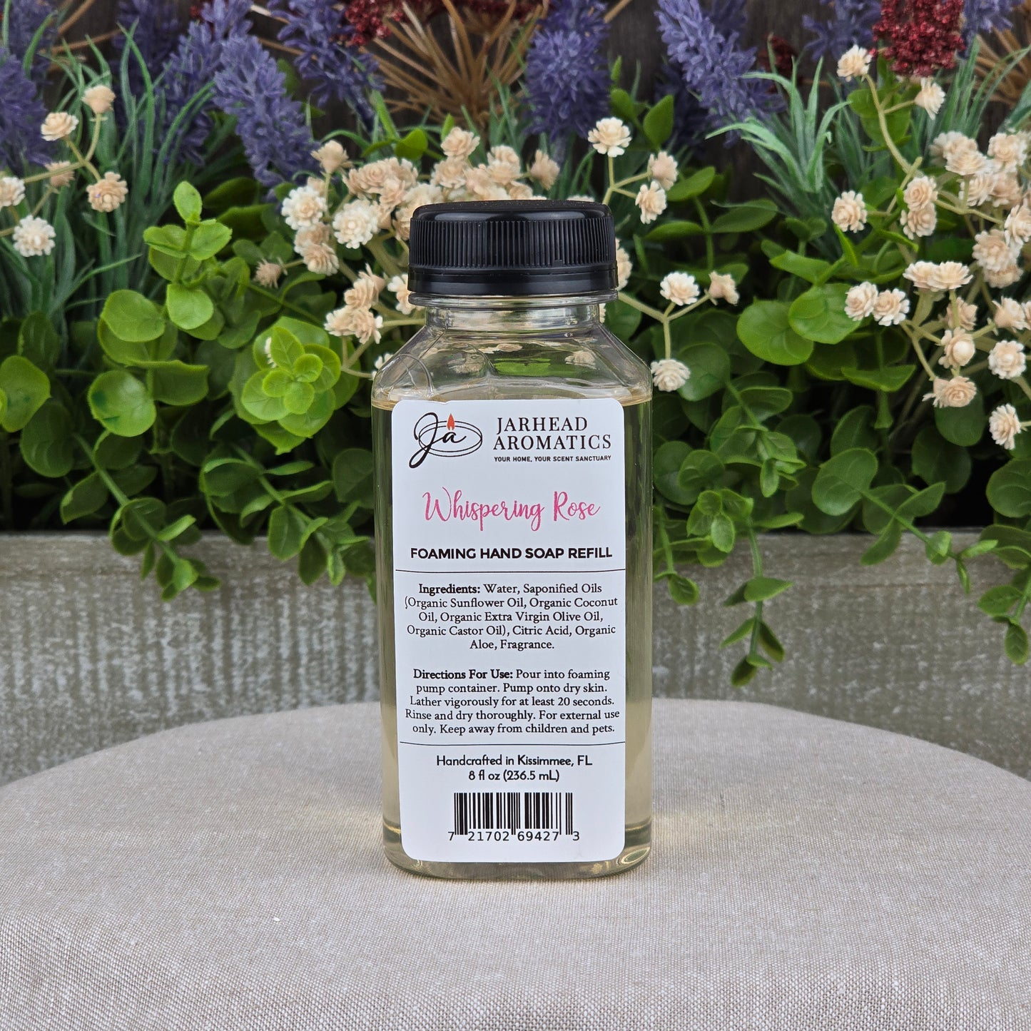 A bottle of Jarhead Aromatics Whispering Rose Foaming Hand Soap rests on a round surface amid blooming flowers and greenery, emphasizing its botanical luxury and plant-based oils.