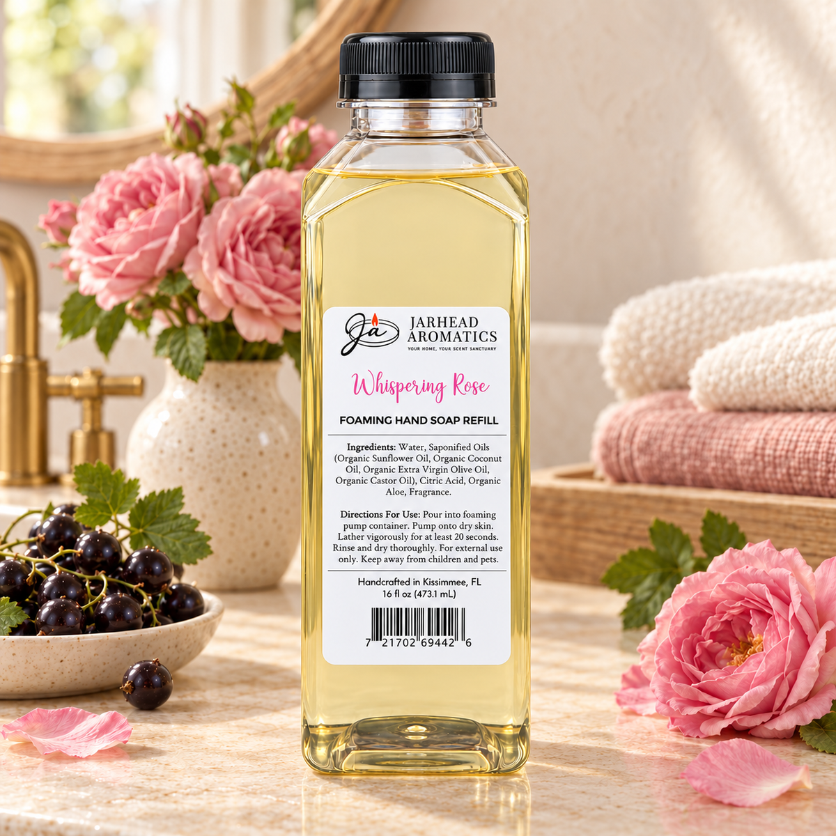 A clear bottle of Jarhead Aromatics Whispering Rose Handmade Foaming Hand Soap sitting on a countertop next to a bowl of black currants and pink roses.