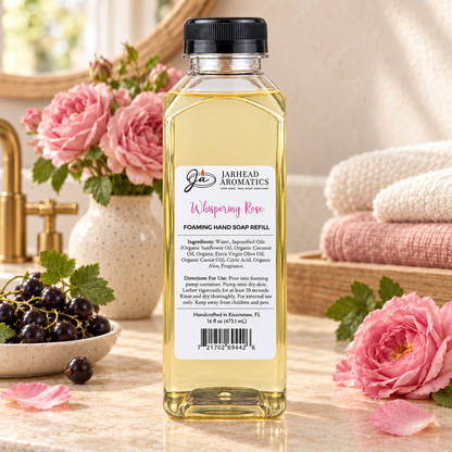 A clear bottle of Jarhead Aromatics Whispering Rose Handmade Foaming Hand Soap sitting on a countertop next to a bowl of black currants and pink roses.