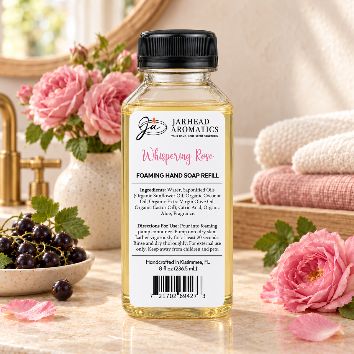 A bottle of Whispering Rose Handmade Foaming Hand Soap | Black Currant, Turkish Rose & White Musk sitting on a counter, surrounded by a bowl of black currants and fresh pink roses.