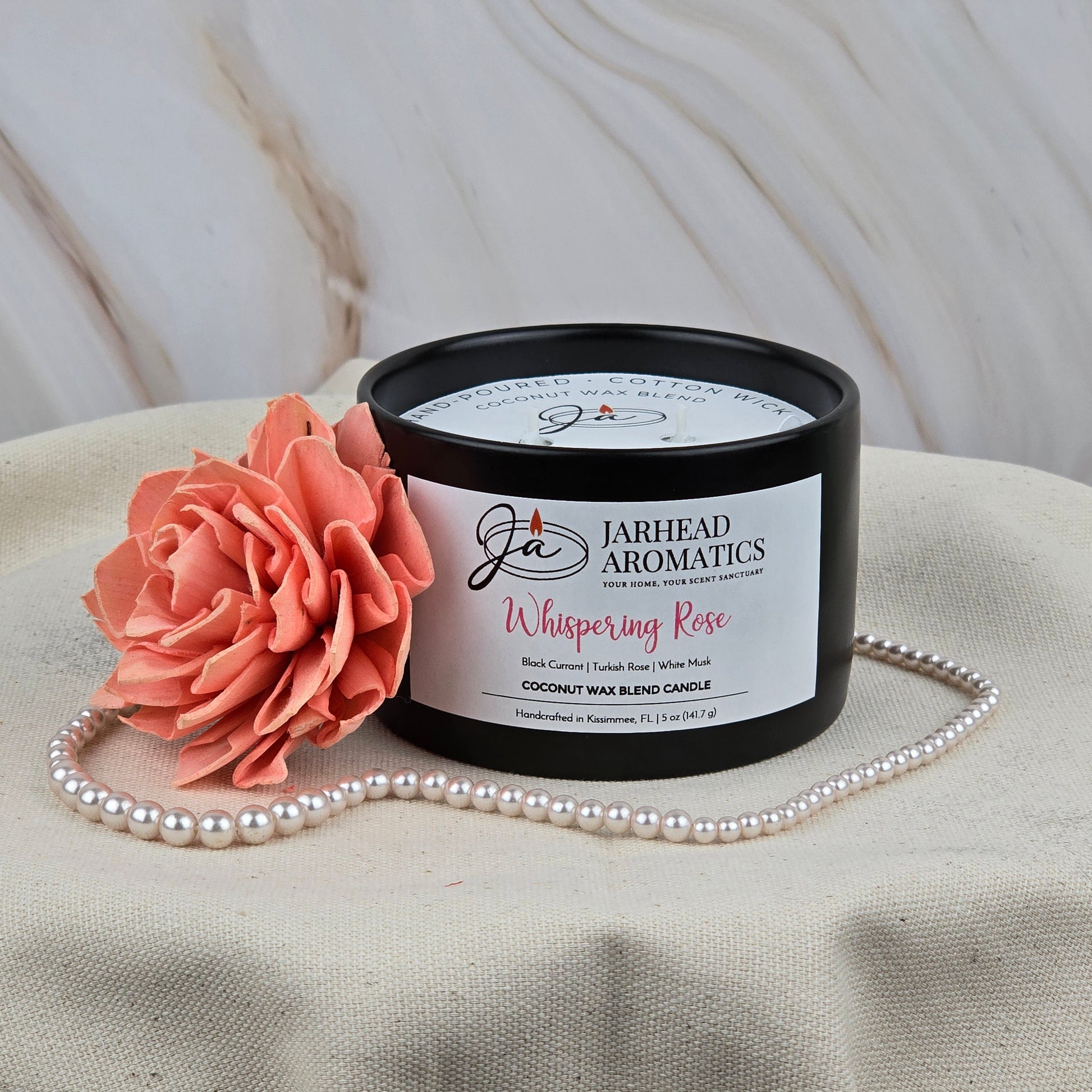 A black Jarhead Aromatics Whispering Rose Coconut Wax Blend Candle rests on beige fabric with a pink artificial flower and pearls, its marble backdrop accentuating the rose, white musk & cedar scent and elegant coconut wax blend.