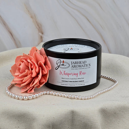 A black Jarhead Aromatics Whispering Rose Coconut Wax Blend Candle rests on beige fabric with a pink artificial flower and pearls, its marble backdrop accentuating the rose, white musk & cedar scent and elegant coconut wax blend.
