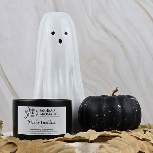 A white ceramic ghost, the Witches Cauldron Coconut Wax Blend Candle by Jarhead Aromatics, and a black pumpkin are displayed among tan faux leaves against a marbled background.