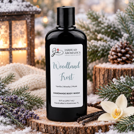 A black bottle of Woodland Frost Handmade Body Wash by Jarhead Aromatics sits on a wooden base surrounded by pine branches, a vanilla flower, and sprigs of lavender.