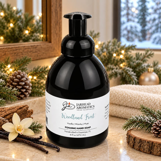 Jarhead Aromatics Woodland Frost Handmade Foaming Hand Soap in a black dispenser with a white label, sitting on a bathroom counter decorated with pine branches, pinecones, and a vanilla bean.