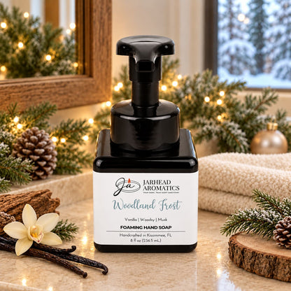 Square bottle of Jarhead Aromatics Woodland Frost Handmade Foaming Hand Soap with a black pump, sitting on a counter with pine branches, pinecones, and a vanilla flower.