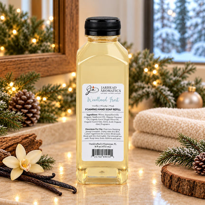 A clear bottle of Woodland Frost Handmade Foaming Hand Soap | Vanilla, Lavender & Woodsy Musk by Jarhead Aromatics sits on a counter with pine branches, pinecones, and a vanilla flower.