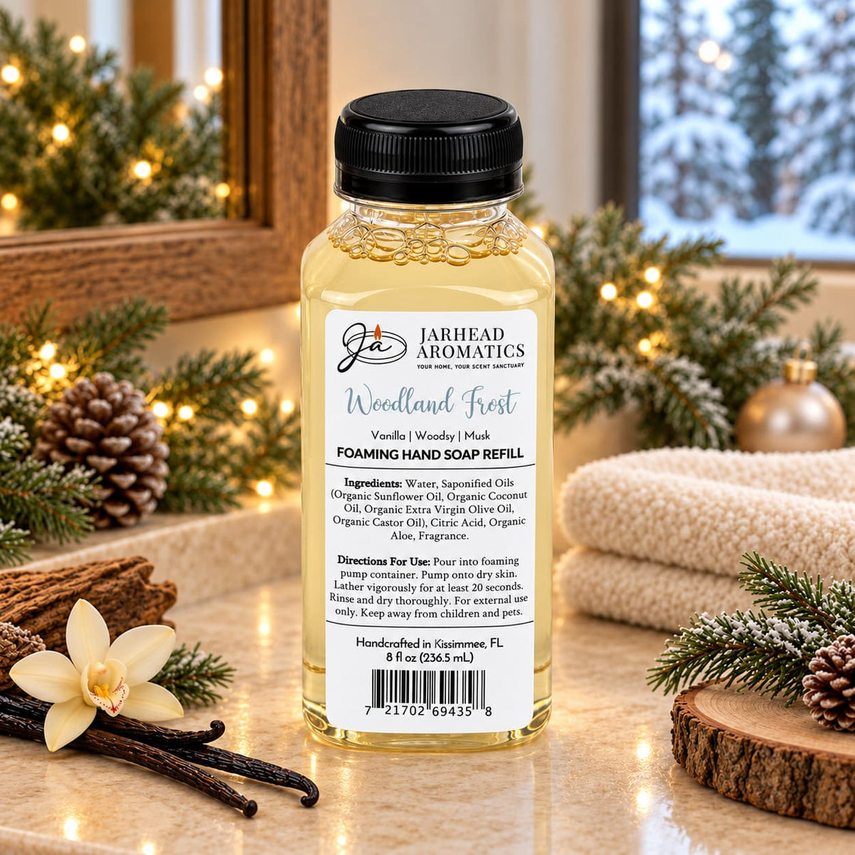 Clear plastic bottle of Jarhead Aromatics Woodland Frost Handmade Foaming Hand Soap | Vanilla, Lavender & Woodsy Musk with an ingredient label, sitting on a counter surrounded by pine branches, pinecones, and a vanilla bean.