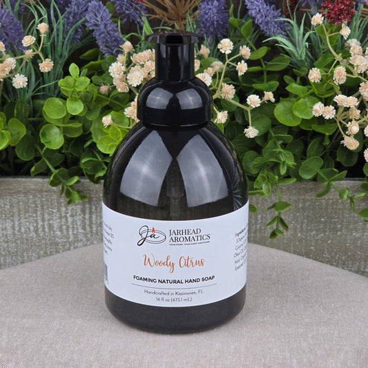A bottle of Jarhead Aromatics Woody Citrus Foaming Natural Hand Soap rests on a gray surface, with artificial greenery and purple and red flowers arranged in the background.
