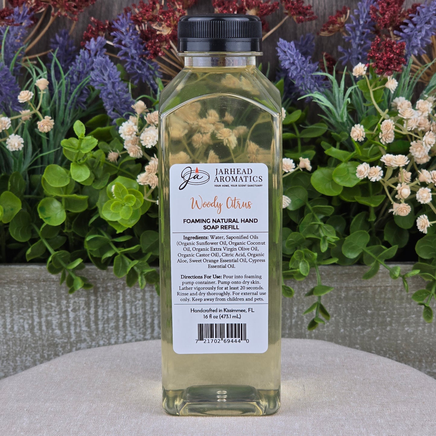A clear plastic bottle of Jarhead Aromatics Woody Citrus Foaming Natural Hand Soap rests on a stone surface, surrounded by green plants and purple flowers.