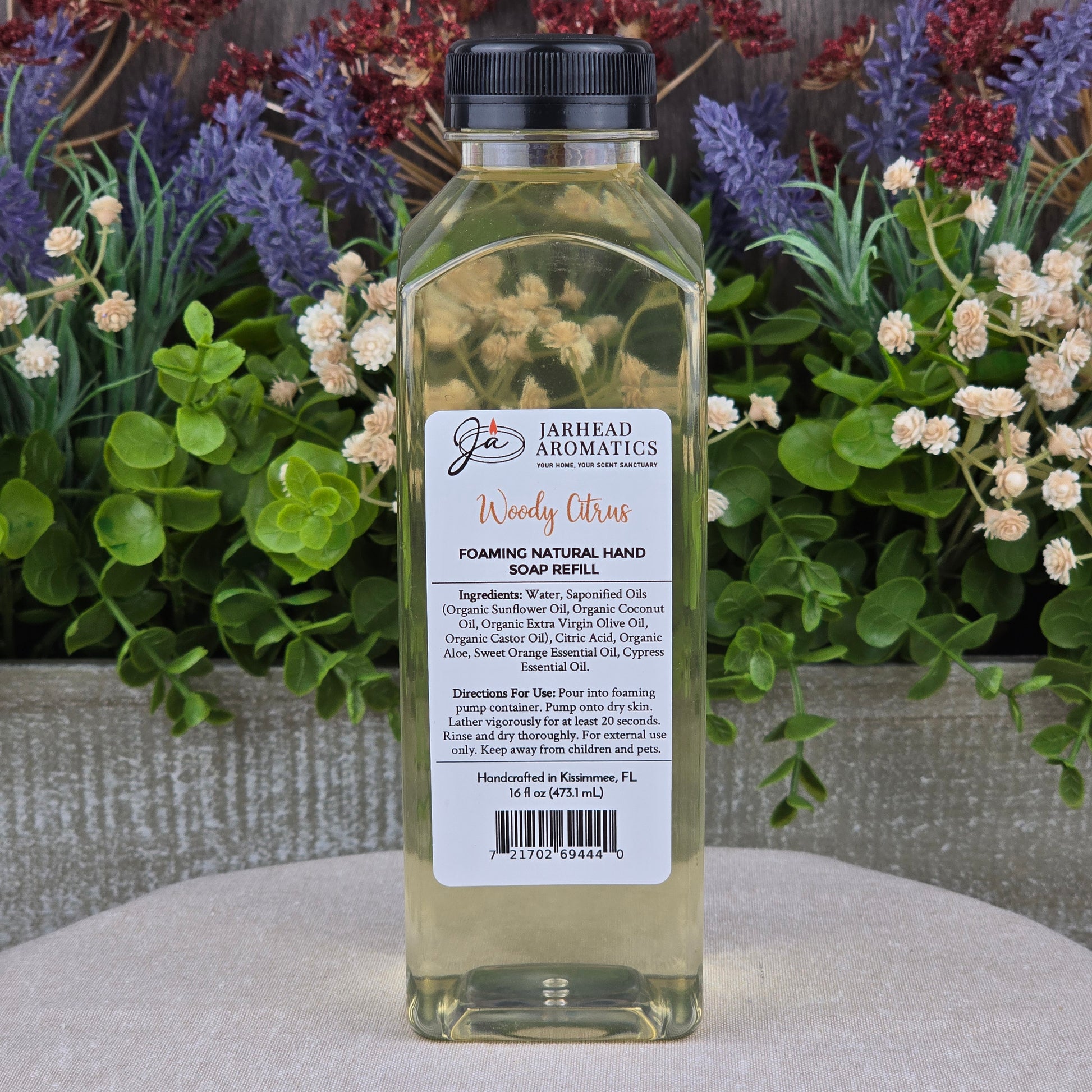 A clear plastic bottle of Jarhead Aromatics Woody Citrus Foaming Natural Hand Soap rests on a stone surface, surrounded by green plants and purple flowers.
