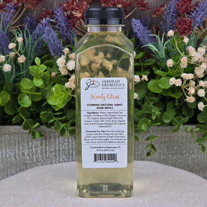 A clear plastic bottle of Jarhead Aromatics Woody Citrus Foaming Natural Hand Soap rests on a stone surface, surrounded by green plants and purple flowers.