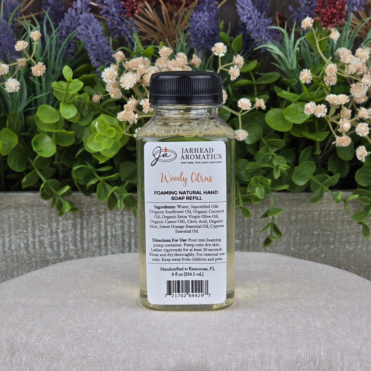 A clear bottle of Jarhead Aromatics Woody Citrus Foaming Natural Hand Soap, a plant-based cleanser with organic oils, sits on a gray surface with its label visible amid green foliage and purple and white flowers.