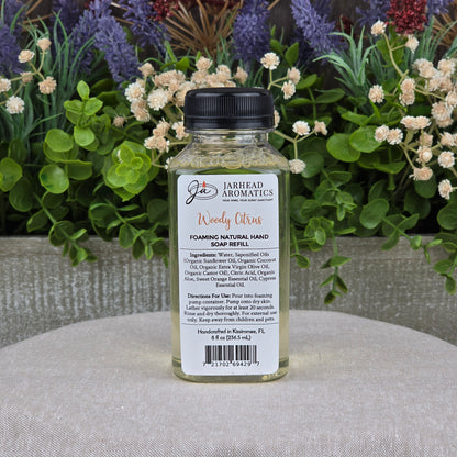 A clear bottle of Jarhead Aromatics Woody Citrus Foaming Natural Hand Soap, a plant-based cleanser with organic oils, sits on a gray surface with its label visible amid green foliage and purple and white flowers.
