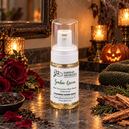 A clear bottle of Zombie Queen Handmade Foaming Hand Soap | Black Tea, Rose Petals & Cedarwood by Jarhead Aromatics sits on a dark countertop, surrounded by red roses, loose tea leaves, and cedar wood chips.