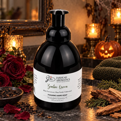 A bottle of Zombie Queen Handmade Foaming Hand Soap with a black pump and white label, surrounded by red roses, loose tea leaves, and wood chips on a dark surface.