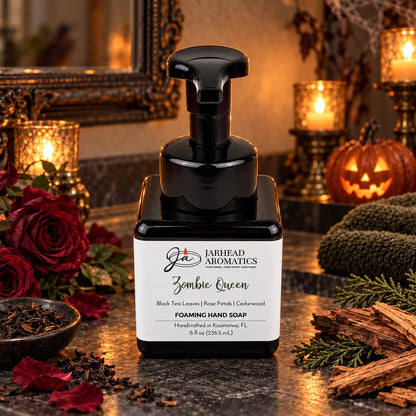 A square bottle of Zombie Queen Handmade Foaming Hand Soap with a black pump dispenser, surrounded by loose tea leaves, rose petals, cedar sticks, and a glowing jack-o-lantern.