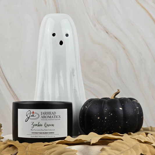 A white ghost figurine, a black pumpkin, and the Jarhead Aromatics Zombie Queen - Coconut Wax Blend Candle are arranged on beige fabric with dried oak leaves, set against a marble-patterned background.